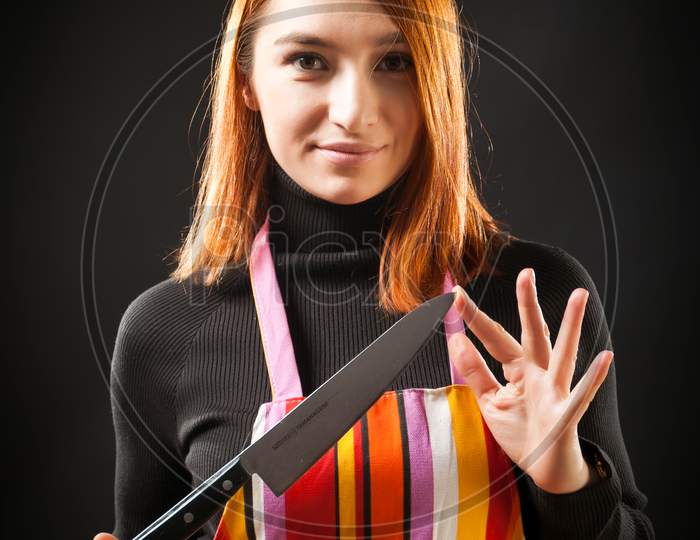 Young Red-Haired Woman In Black Sweater And Kitchen Apron Holds A Kitchen Knife In Her Hand, Shows It At The Camera And Smiles On A Black Isolated Background