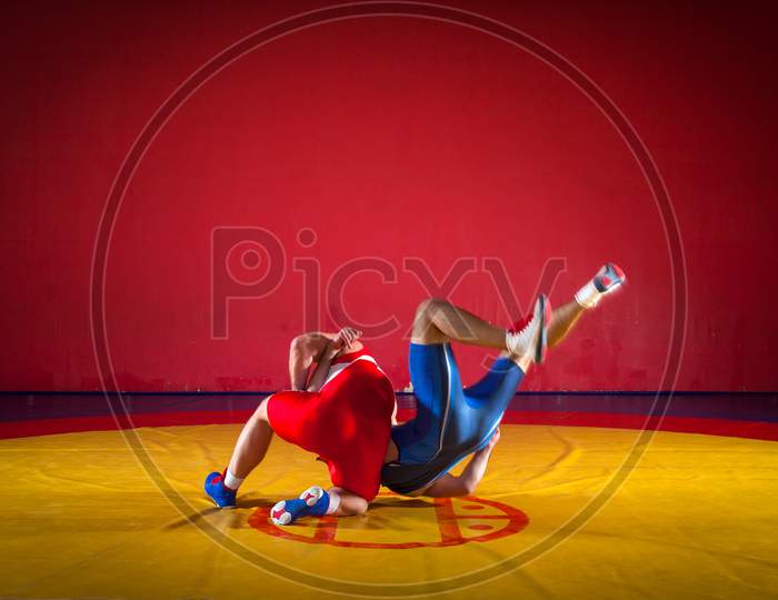 Two Strong Wrestlers In Blue And Red Wrestling Tights Are Wrestlng And Making A Suplex Wrestling On A Yellow Wrestling Carpet In The Gym. Young Man Doing Grapple.