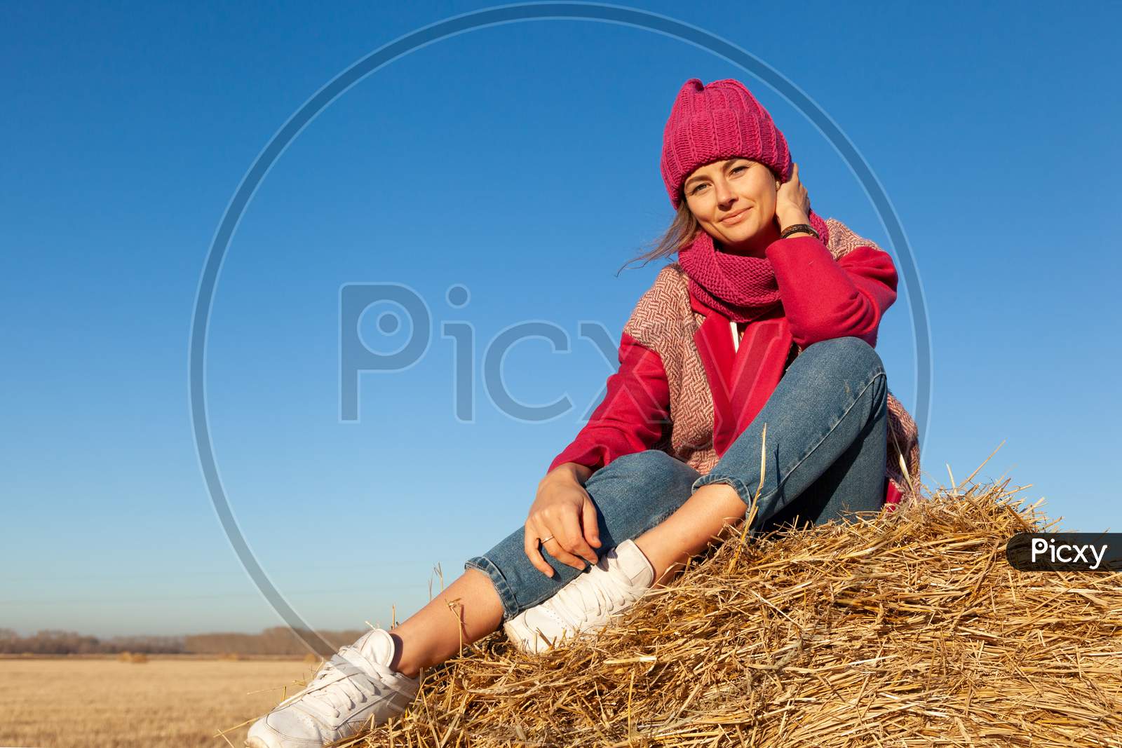 The Concept Of Livestyle  Outdoor In Autumn. Close Up Of A Young Woman Student In A Warm Autumn Clothes Looking Funny, Smilling, Posing For The Camera, Sitting On Haystick