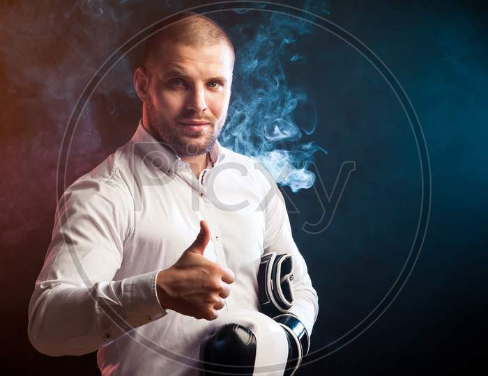 Handsome Young Man Businessman In White Shirt And Black And White Boxing Gloves Smiles And Shows Thumb Up Against Red And Blue Smoke From A Wipe On A Black Isolated Background