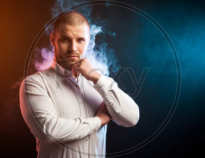Handsome Man Sportsman In A White Shirt Stands With Arms Crossed On His Chest And Confidently Looks In The Camera Opposite Red And Blue Smoke From A Wipe On A Black Isolated Background