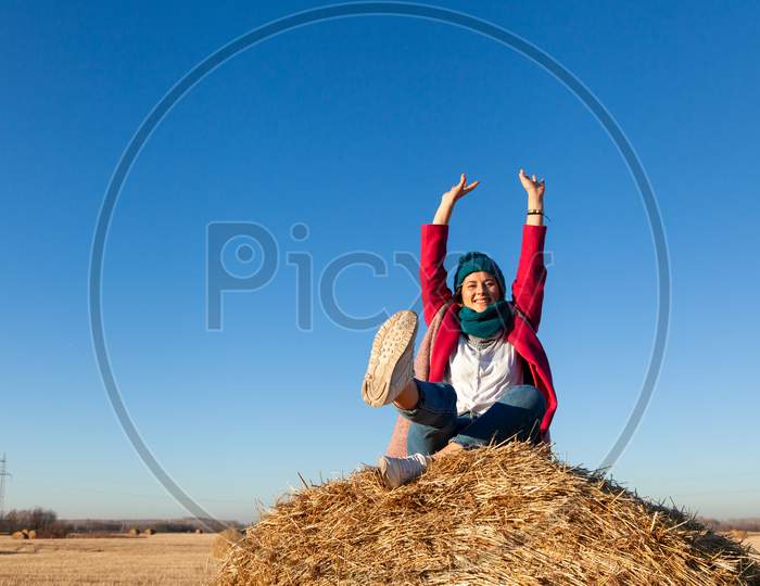 Portrait Female Student On Field Background. Beautiful Modern  Young Woman Wearing Pink Knitting Hat  Smile, Look At The Camera And Enjoy Autumn Sunny Day And Sitting On Haystick.
