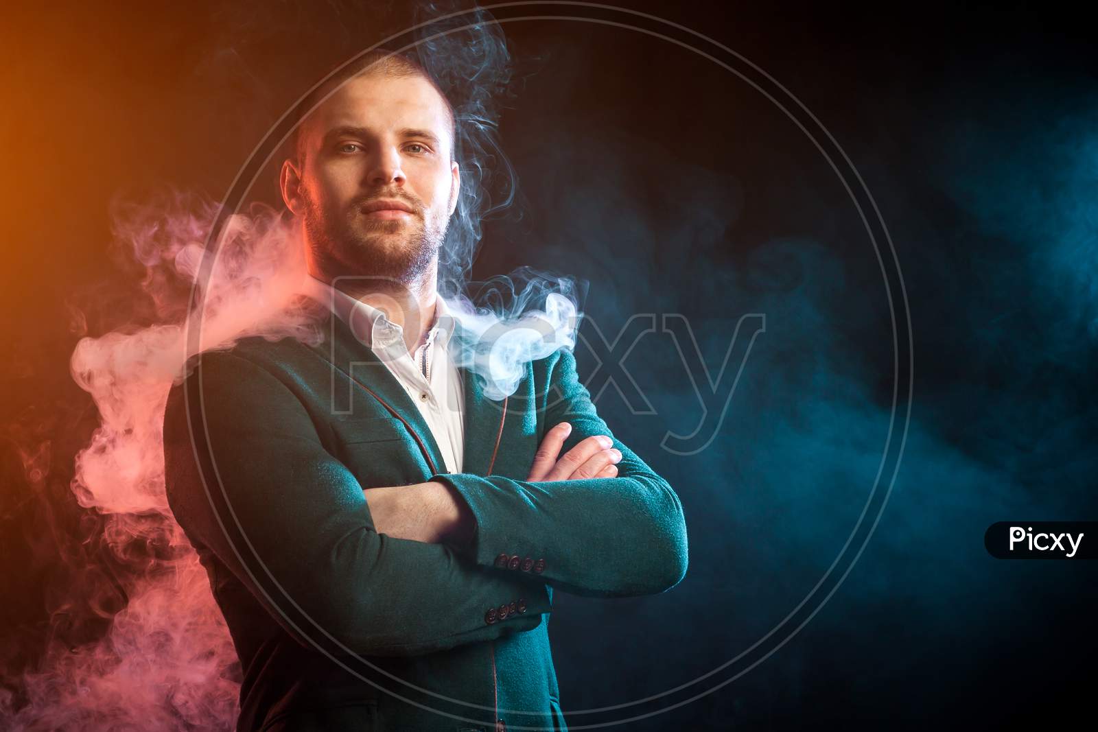 Young Handsome Man Head In A White Shirt And Green Jacket Smiling And Looking Thoughtfully Into The Camera Opposite Red And Blue Smoke From A Wipe On A Black Isolated Background