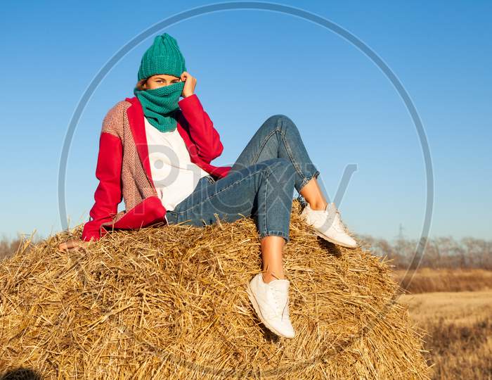 Outdoor Atmospheric Lifestyle Photo Of Young Beautiful Lady With Brown Hair And Eyes In Pink Coat, Knitting Hat And Jeans Posing And Sitting On Hay Around  Blue Sky. Warm Autumn.