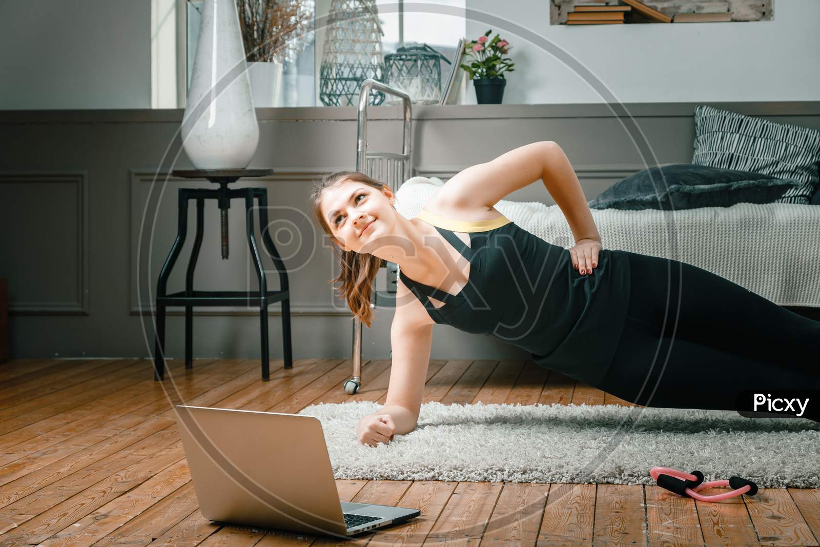 The Young Woman Goes In For Sports At Home. Sportsman With Black Hair Makes A Plank, Watches A Movie And Studies From A Laptop  On Carpet In Bedroom