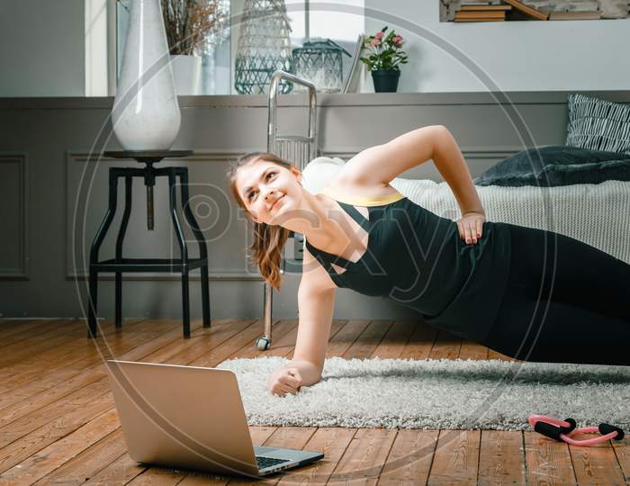 The Young Woman Goes In For Sports At Home. Sportsman With Black Hair Makes A Plank, Watches A Movie And Studies From A Laptop  On Carpet In Bedroom