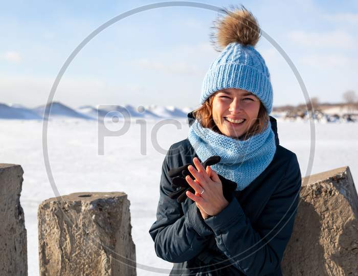 Portrait Beauty Woman Model On Winter Background. Beautiful Modern Urban Young Woman Wearing Blue Knitting Hat  Basks In Cold Weather On The Background Of The Frozen Sea