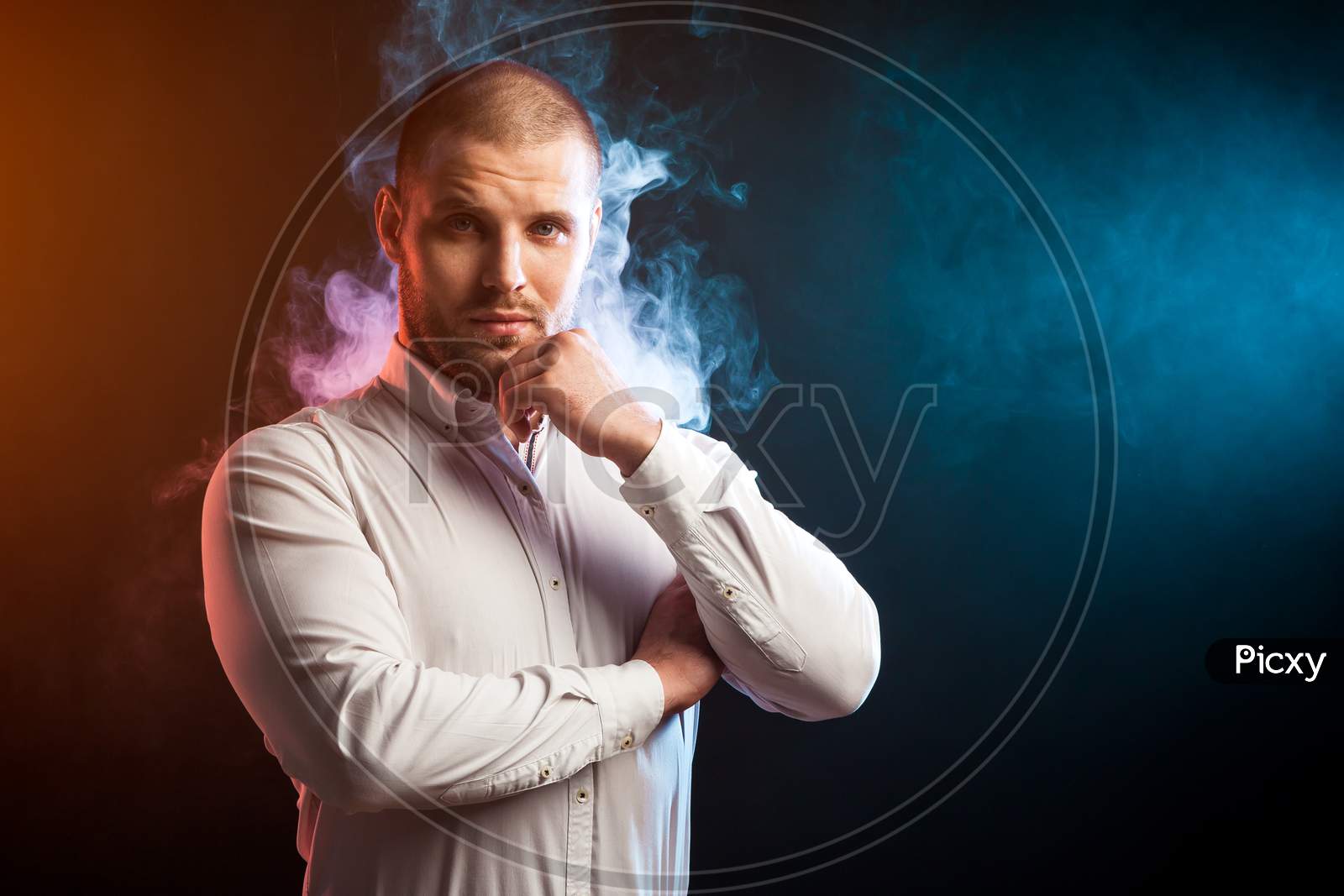 Handsome Man Sportsman In A White Shirt Stands With Arms Crossed On His Chest And Confidently Looks In The Camera Opposite Red And Blue Smoke From A Wipe On A Black Isolated Background