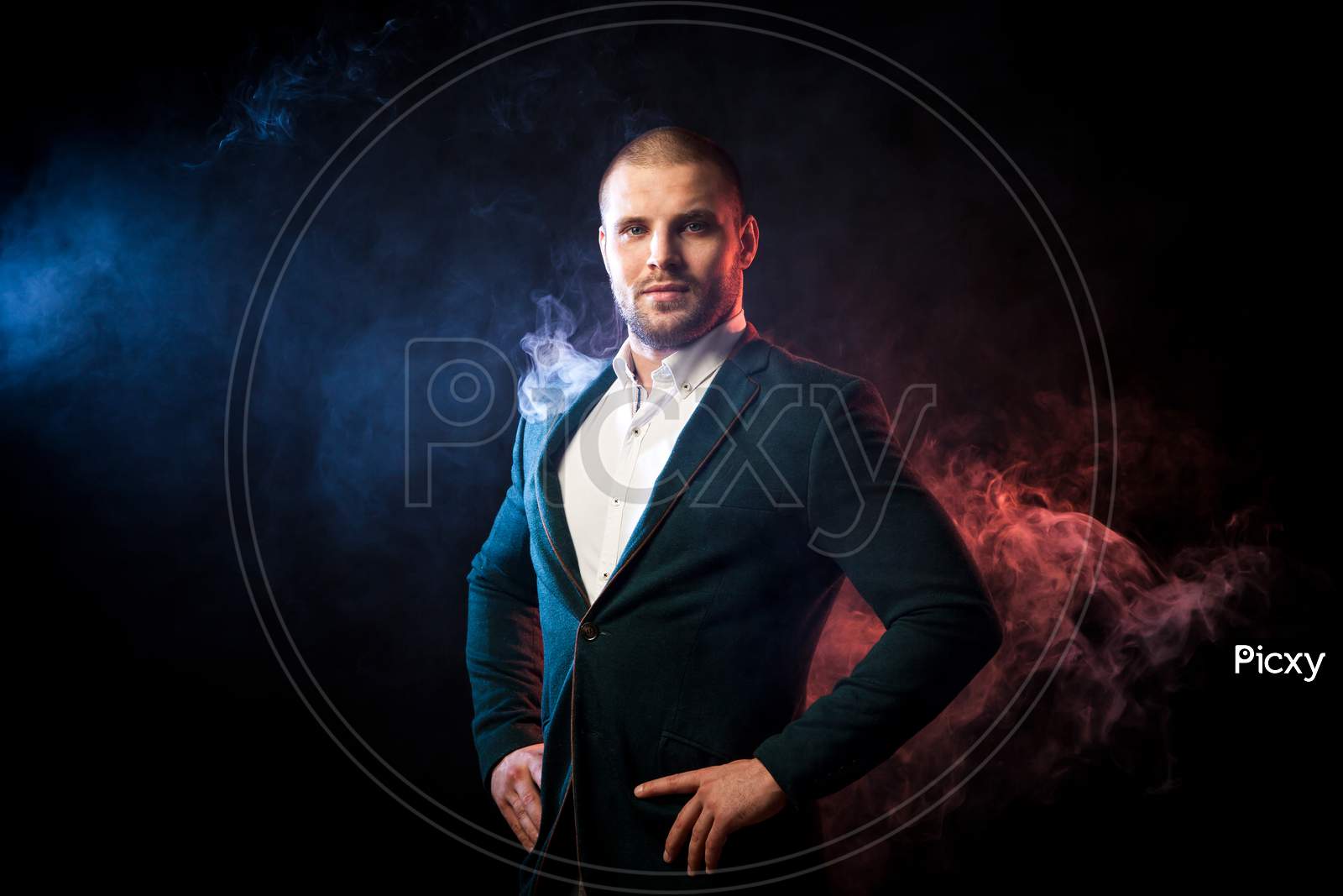 Young Handsome Man Manager In White Shirt And Green Jacket Posing Against Red And Blue Smoke From A Wipe On Black Isolated...