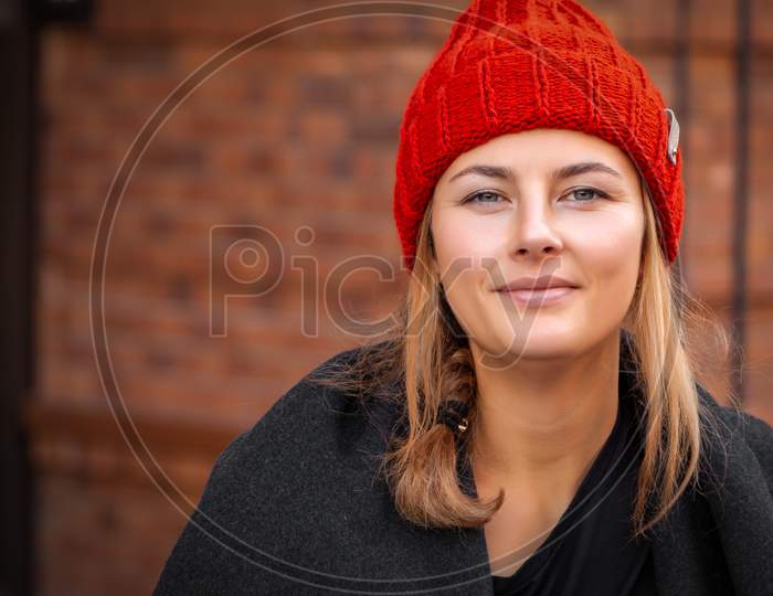 Fashion Lifestyle Portrait Of Young Trendy Woman Dressed In Black Coat And  Red Knitted Hat Sits On Bench, Look At The Camera, Enjoy Vacation  In The  Old City, Autumn Street Fashion.  Portrait Of Joyful Woman