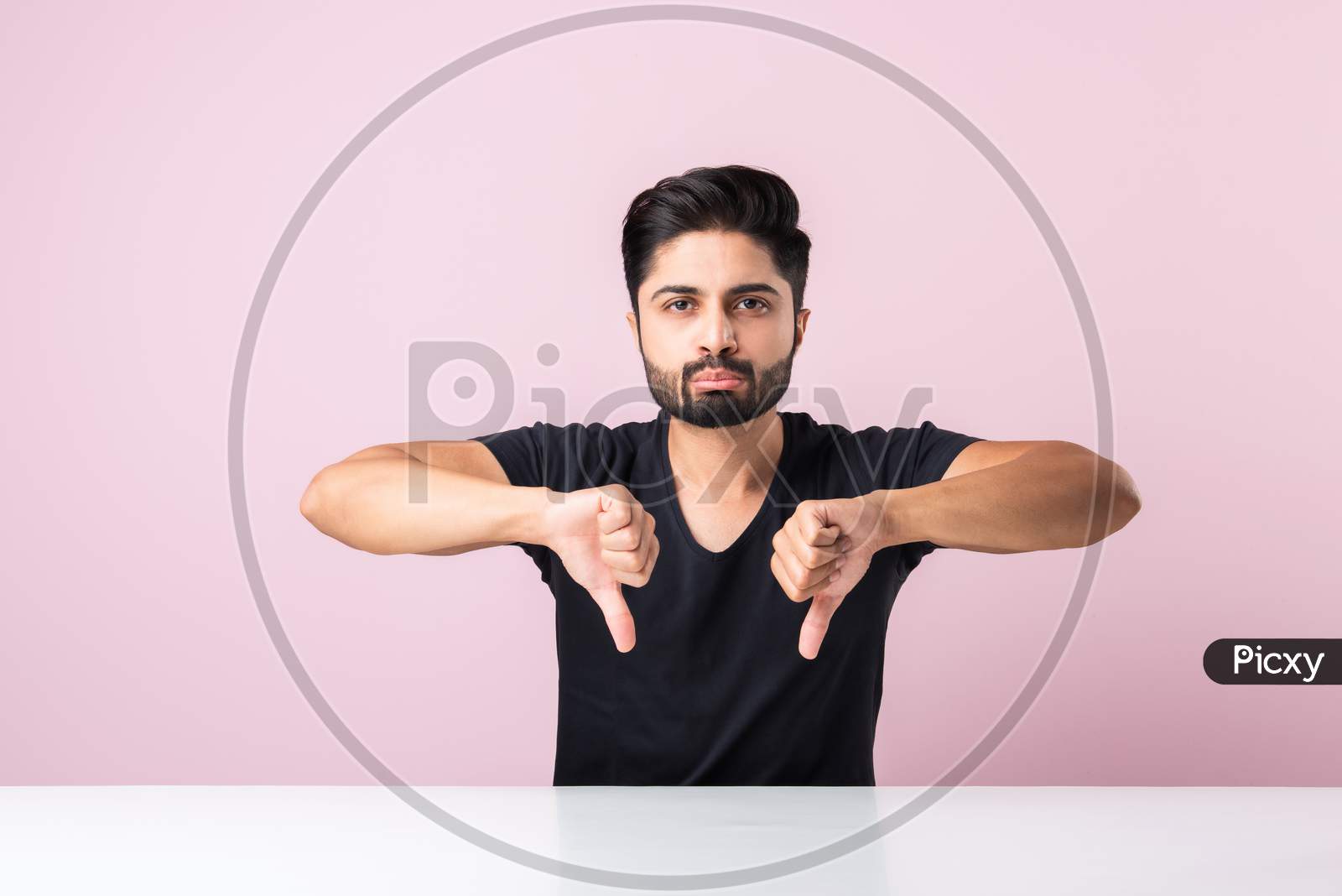 Indian Asian Young Man With Negative Emotions Showing Thumbs Down Symbol, Sitting At Table