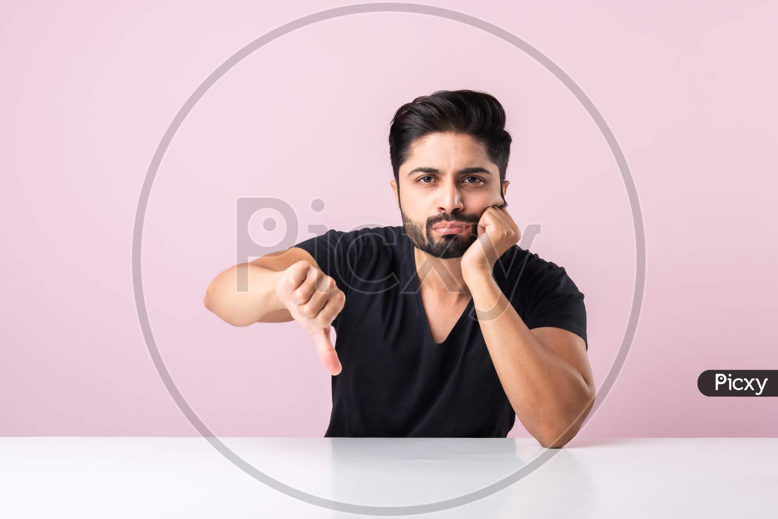 Indian Asian Young Man With Negative Emotions Showing Thumbs Down Symbol, Sitting At Table