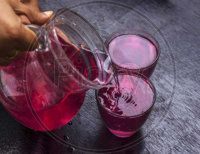 A Female Hand Pouring Red Drink To Two Glasses From A Jug.