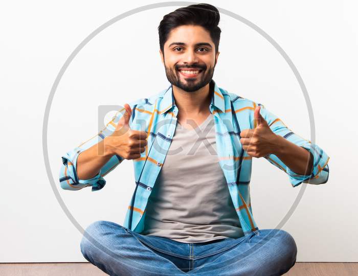 Indian Bearded Young Man With Thumbs Up Or Success Or Ok Symbol Sitting Isolated Over Wooden Floor