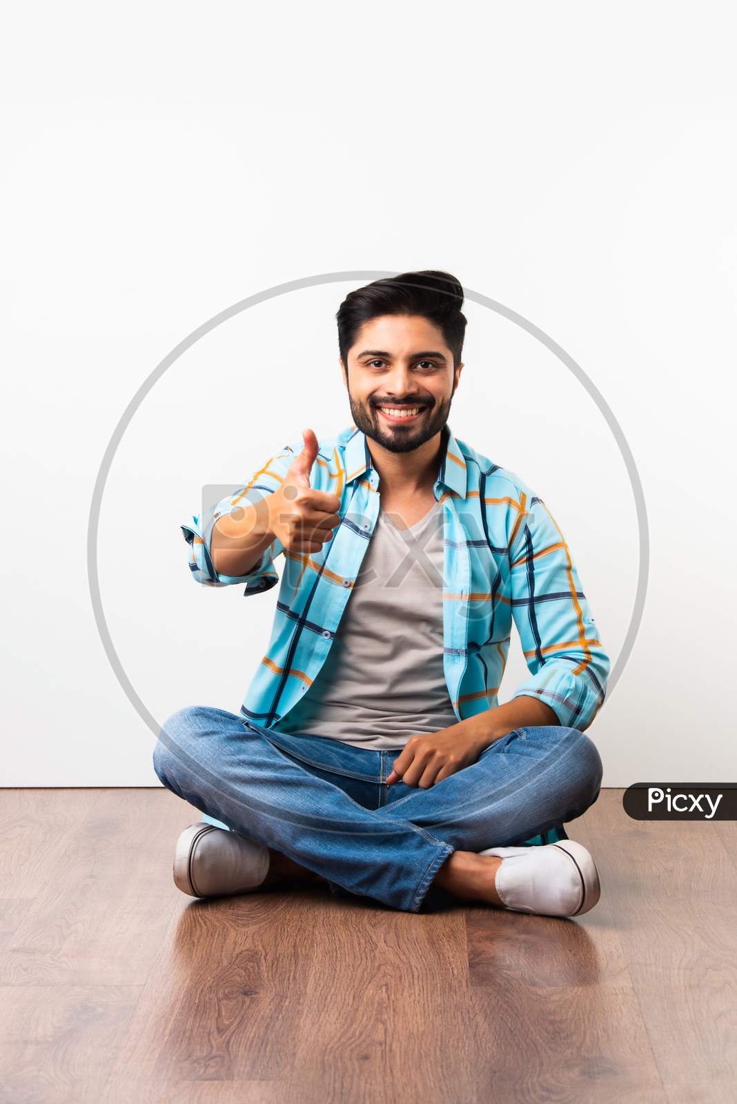 Indian Bearded Young Man With Thumbs Up Or Success Or Ok Symbol Sitting Isolated Over Wooden Floor