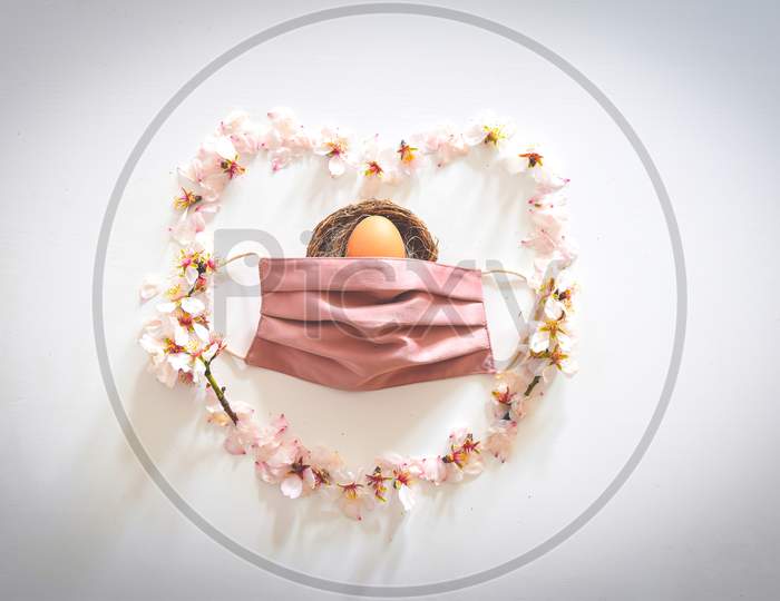 Spring Daisy Flowers Forms Heart With Pink Facial Mask In Middle Cover Easter Egg. Conceptual Safe Easter Anti-Pandemic White Background