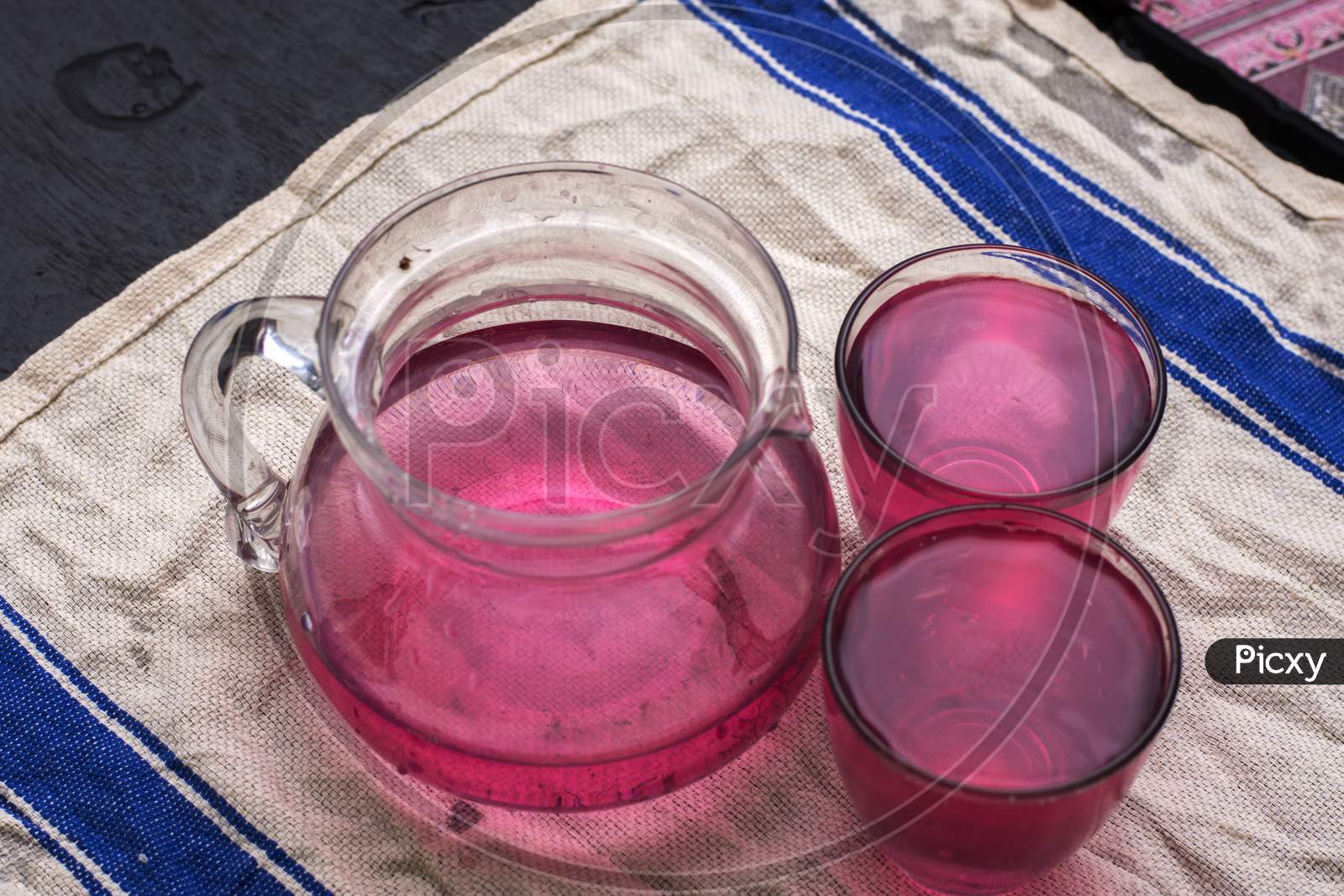A Jug And Two Glasses Full Of Red Drink On A White Mat.