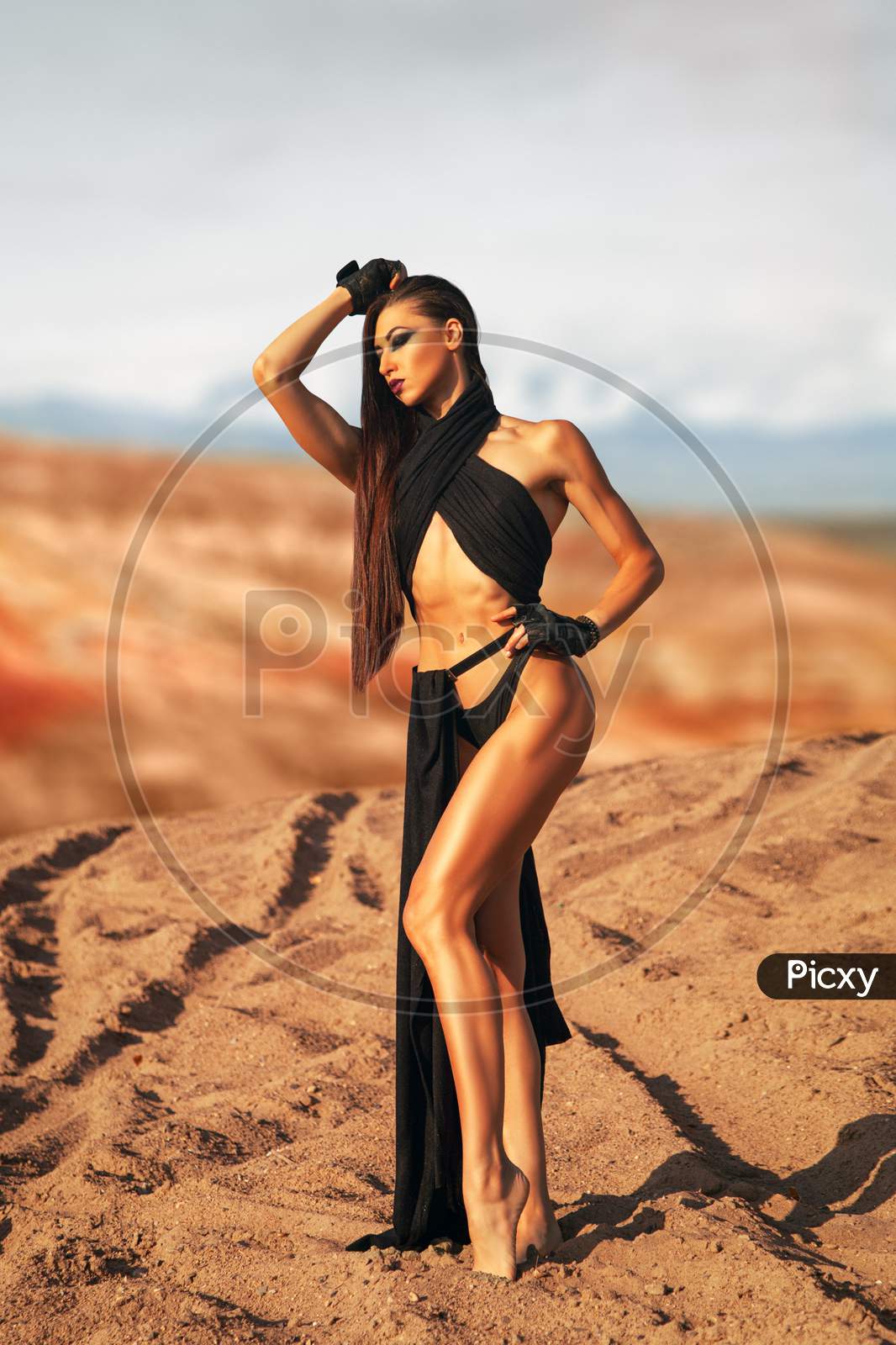 A Young Woman With A Gothic Make-Up In A Black Conceptual Swimsuit In A Gladiatorial Style Is Thoughtfully Posing Against The Backdrop Of Deserts And Mountains.The Concept Of Oriental Tales And Beauty