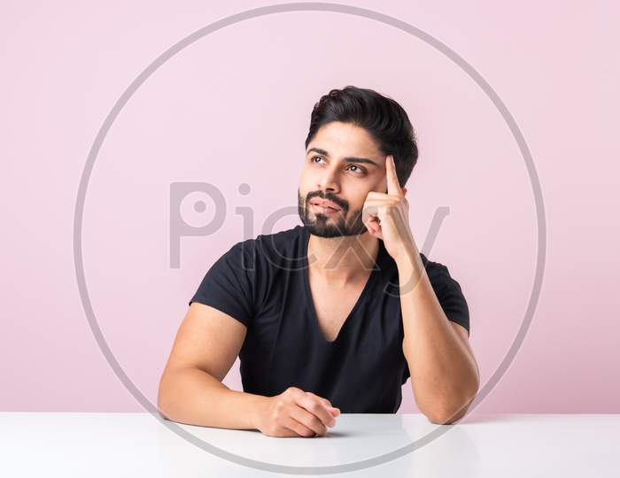 Indian Bearded Young Man Thinking While Sitting Isolated Against Pink Background At Table Or Desk