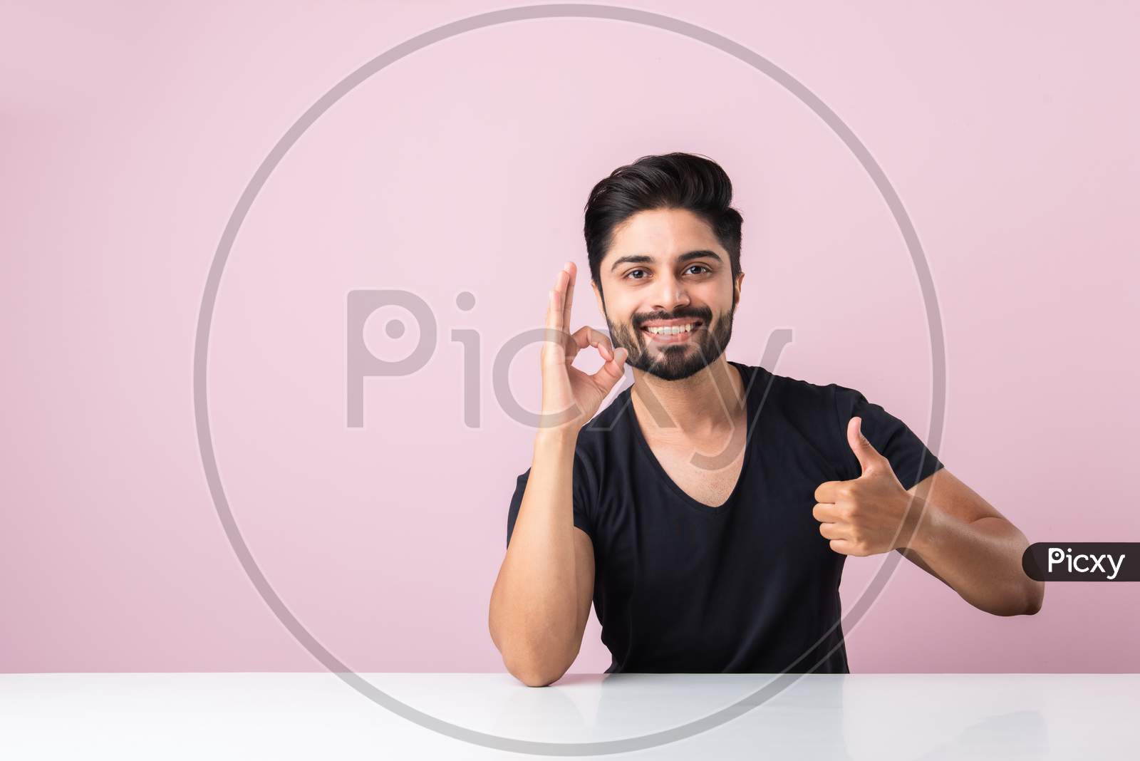 Indian Bearded Young Man With Thumbs Up Or Success Or Ok Symbol Sitting At Table Isolated