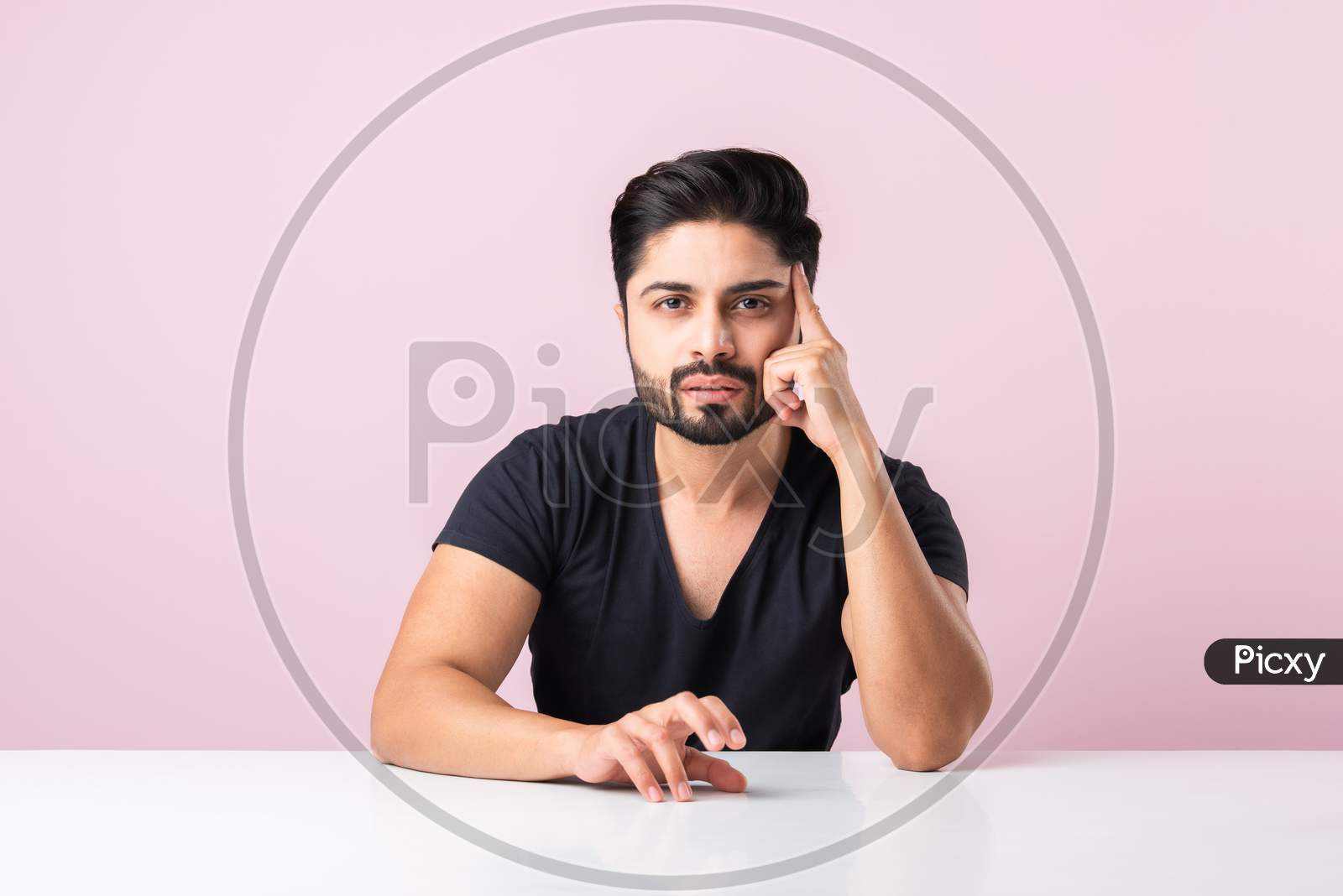 Indian Bearded Young Man Thinking While Sitting Isolated Against Pink Background At Table Or Desk