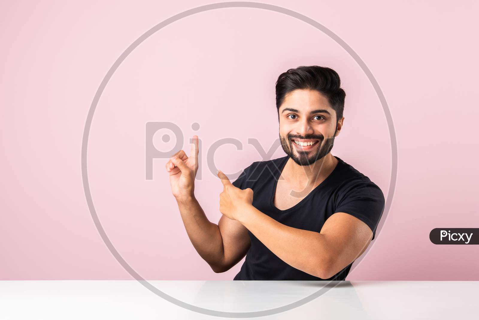 Indian Asian Man Presenting, Pointing Or Holding Something While Sitting Isolated Over Pink At Table