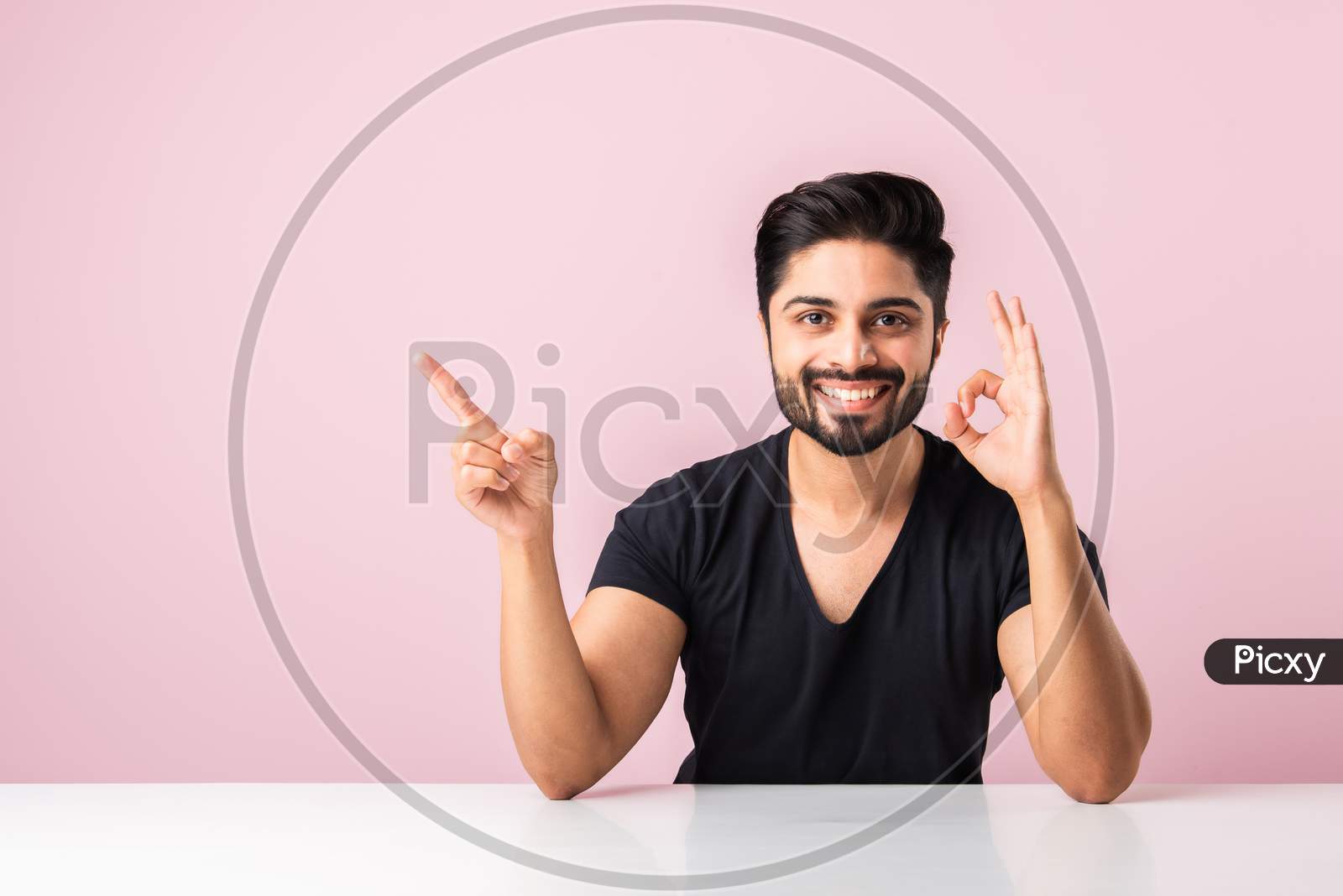 Indian Asian Man Presenting, Pointing Or Holding Something While Sitting Isolated Over Pink At Table