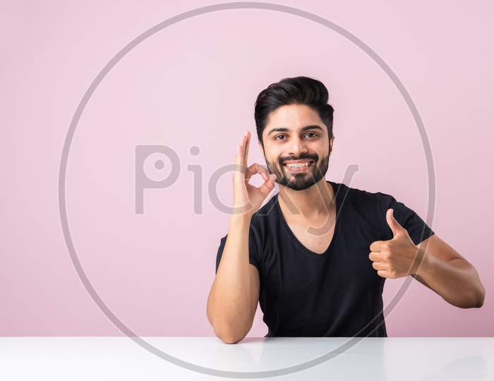 Indian Bearded Young Man With Thumbs Up Or Success Or Ok Symbol Sitting At Table Isolated