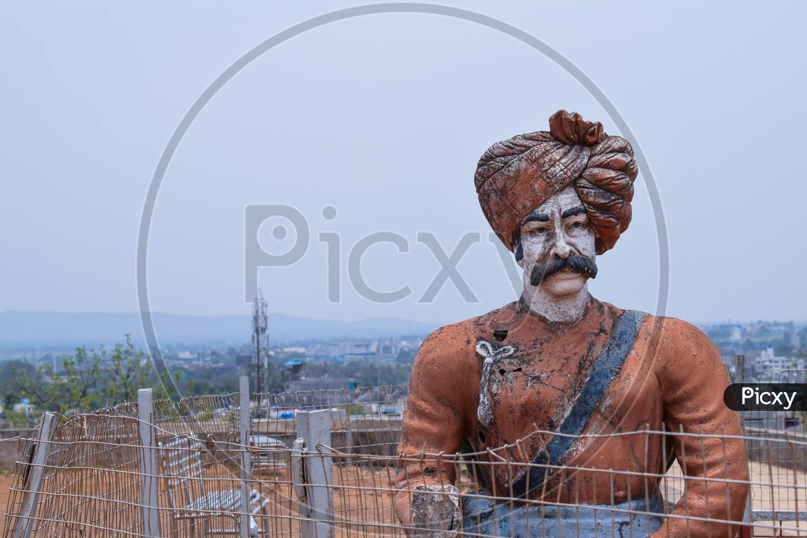 Front View Of 30-40 Years Old Indian Warrior Statue, Wearing Orange Color Turban And Indian Traditional Cloths Standing Still In A Sunny Day At Kolhapur City Maharashtra India On Blur Background With Copy Space.