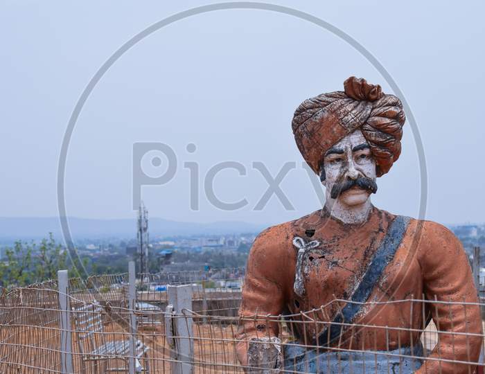Front View Of 30-40 Years Old Indian Warrior Statue, Wearing Orange Color Turban And Indian Traditional Cloths Standing Still In A Sunny Day At Kolhapur City Maharashtra India On Blur Background With Copy Space.
