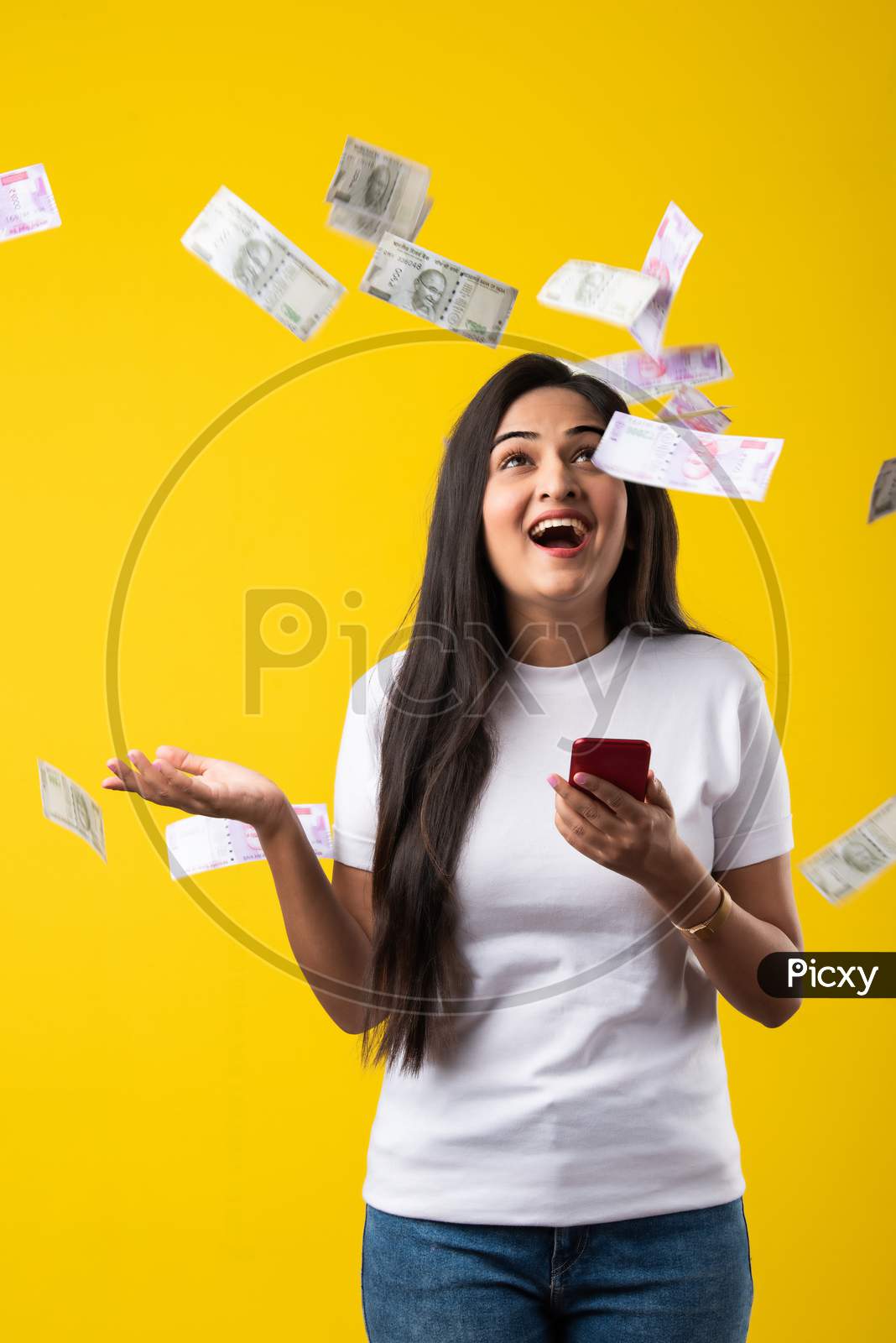 Indian Pretty Woman Using Smartphone While Currency Notes Falling Against Yellow Background