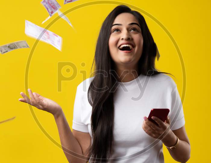 Indian Pretty Woman Using Smartphone While Currency Notes Falling Against Yellow Background