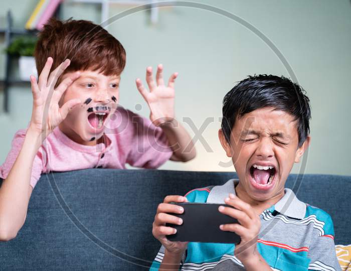 Young Kid With Face Painting Fools Or Making Prank To His Brother While He Is Busy On Mobile By Suddenly Screaming And Shouting From Back Side At Home During April Fools Day Celebration.