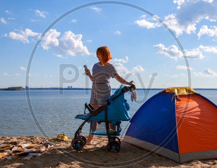 A Young Mother Rolls Her Newborn Baby In A Stroller And Looks Into The Phone On A Warm Summer Day On The Sea With A Sandy Beach. Family Vacation At Sea