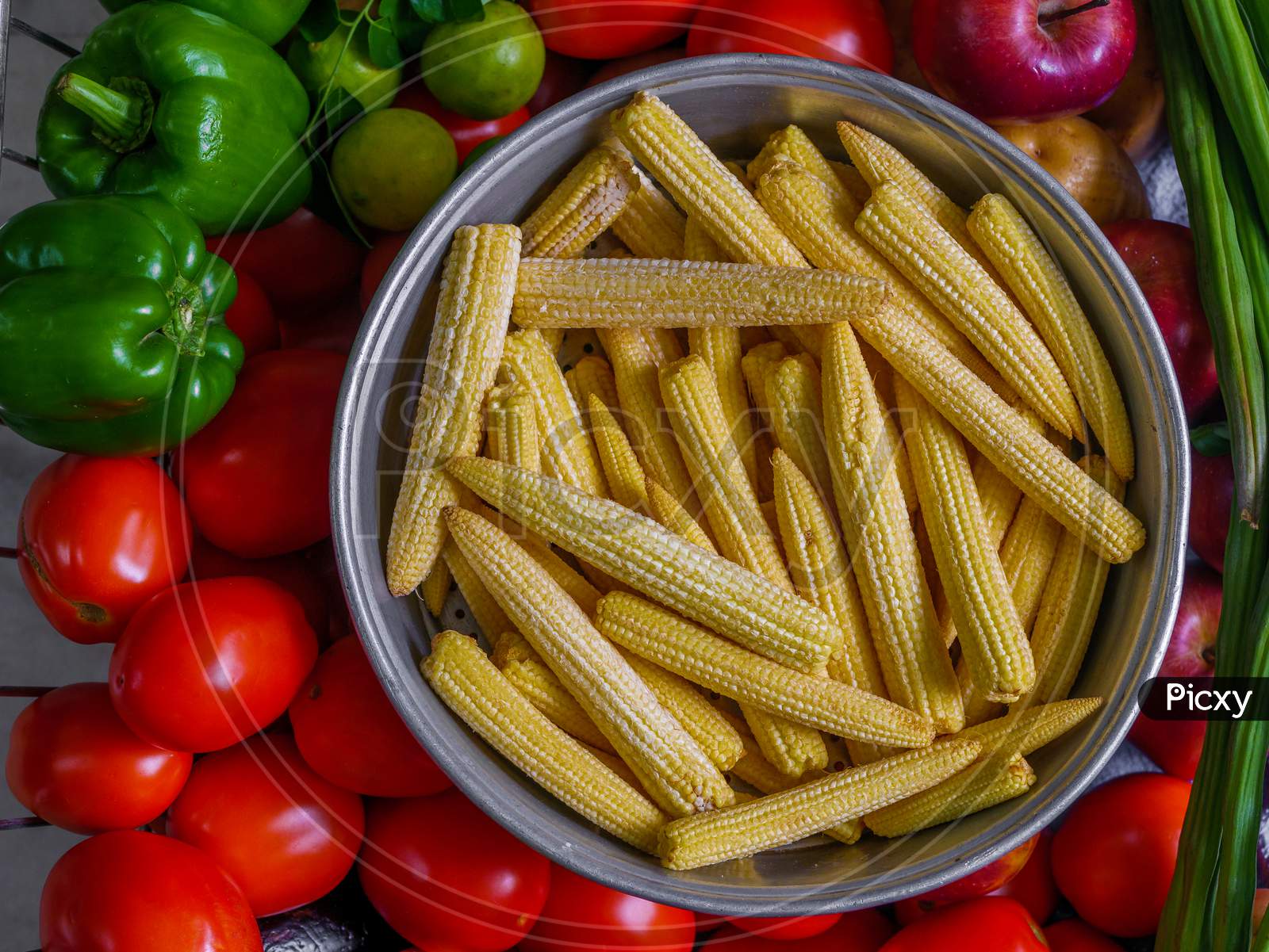 Baby Corns Arranged In A Basket With Tomatoes And Green Veggies In Background