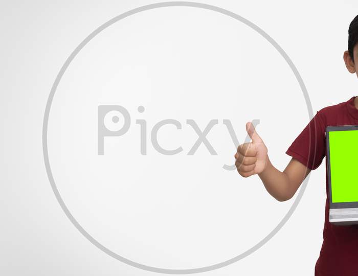 Banner Image Of A Young Indian Kid Holding A Laptop With Green Screen In His Hands And Showing Thumbs Up To The Camera. Standing On A White Isolated Wall With Copy Space.