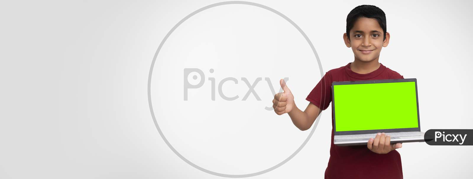 Banner Image Of A Young Indian Kid Holding A Laptop With Green Screen In His Hands And Showing Thumbs Up To The Camera. Standing On A White Isolated Wall With Copy Space.