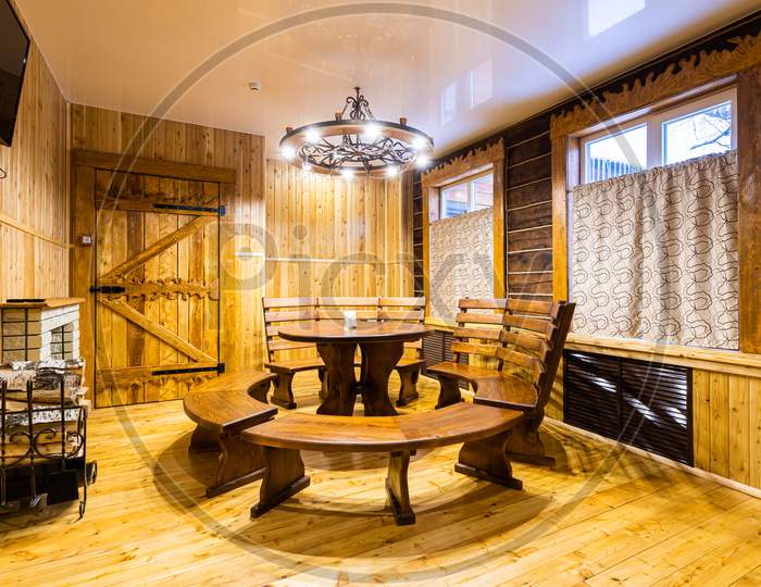 Living Room Of A Country House: Wall, Decorated With Clapboard, Medieval-Style Chandelier, Large Wooden Table With A Bench, Window Frames And A Fireplace With Logs