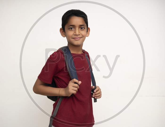 Young Indian Kid Standing On A White Wall With A School Bag On His Back. Getting Ready For School. School And Education Concept.