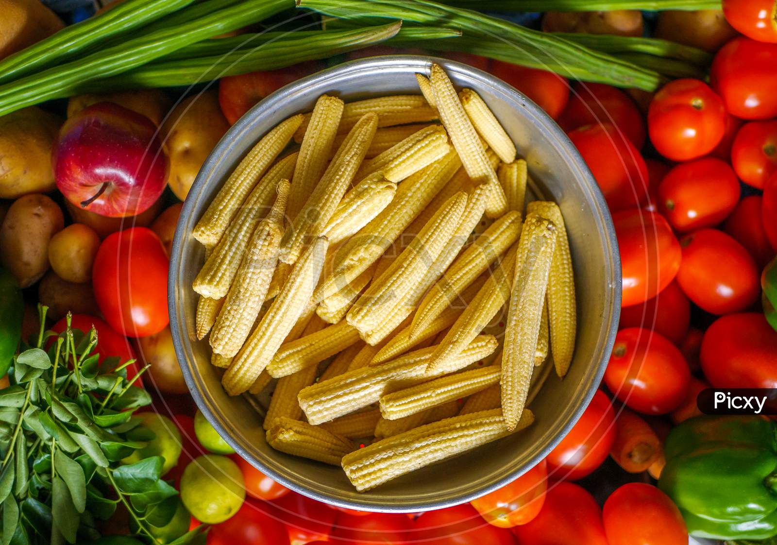 Baby Corns Arranged In A Basket With Tomatoes And Green Veggies In Background