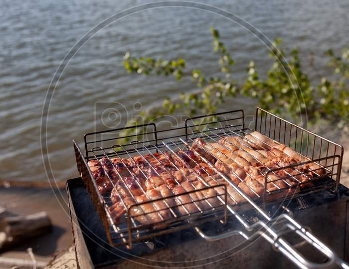 Chicken Meat Is Fried On A Small Folding Grill In A Metal Grill, Against A Background Of Blue Sky, Sun And Sand. The Concept Of Departure For Barbecue And Sea Rest