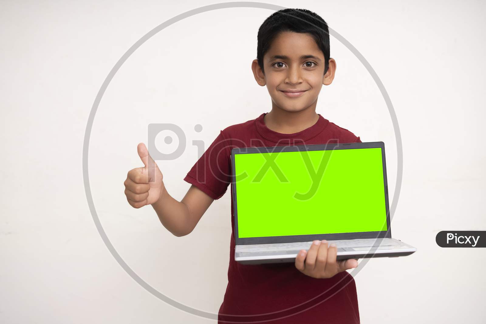Young Indian Kid Holding A Laptop With Green Screen In His Hands And Showing Thumbs Up To The Camera. Standing On A White Isolated Wall With Copy Space.