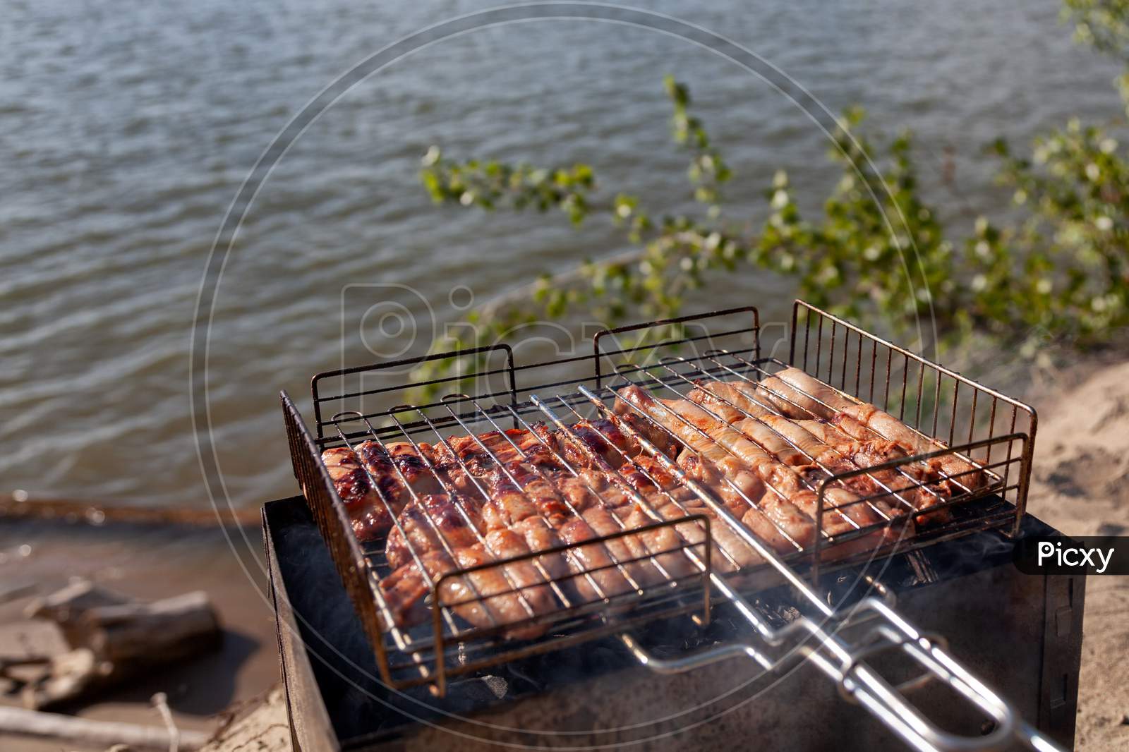 Chicken Meat Is Fried On A Small Folding Grill In A Metal Grill, Against A Background Of Blue Sky, Sun And Sand. The Concept Of Departure For Barbecue And Sea Rest