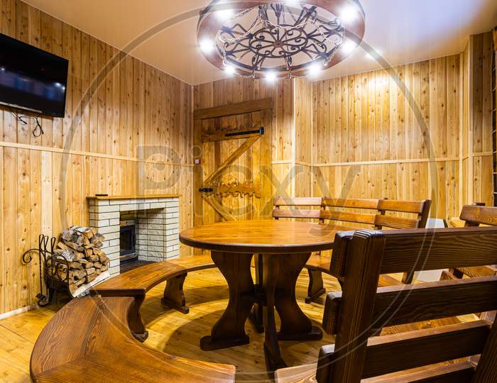 Living Room Of A Country House: Wall, Decorated With Clapboard, Medieval-Style Chandelier, Large Wooden Table With A Bench, Window Frames And A Fireplace With Logs