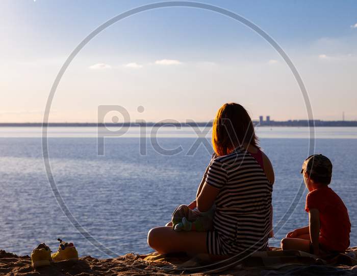 A Young Mother With Her Children: A Boy And A Newborn Are Sitting And Enjoying The Nature Of The Sea, Watching The Sunset On A Warm Summer Day. Family Vacation At Sea