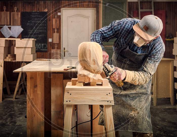 Experienced Carpenter In Work Clothes And Small Buiness Owner Working In Woodwork Workshop, Processes The Board With An Angle Grinder On The Table Is A Hammer And Many Tools