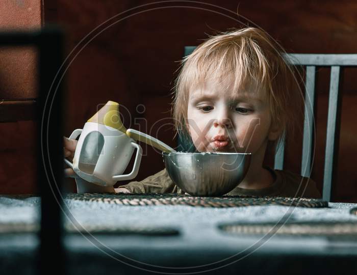 A Small Child Of Two Years Old In The Kitchen Eats Mashed Potatoes And Drinks Water From A Baby Sippy Cup At The Dining Table In The Kitchen