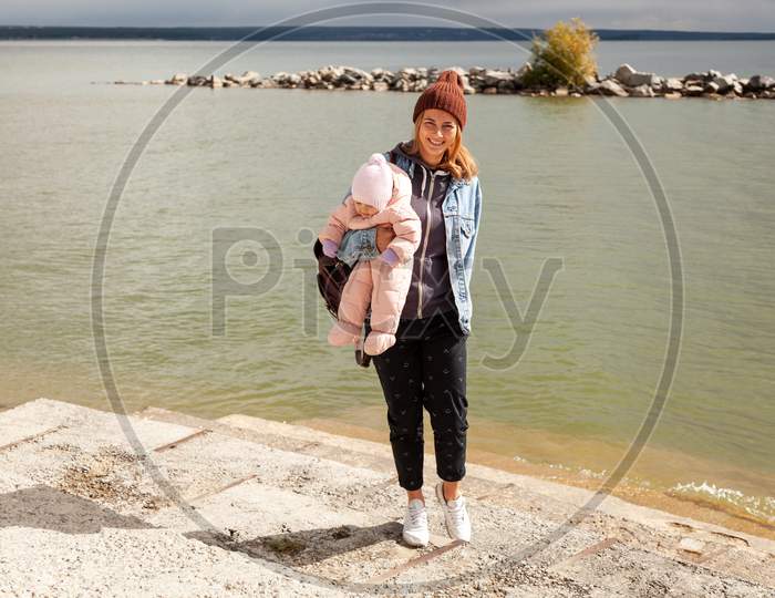 A Young  Mother Playing Her Daughter And Enjoying Nature On The Back Of A Background Of Autumn Sea. The Concept Of Livestyle And Family Outdoor Recreation In Autumn.