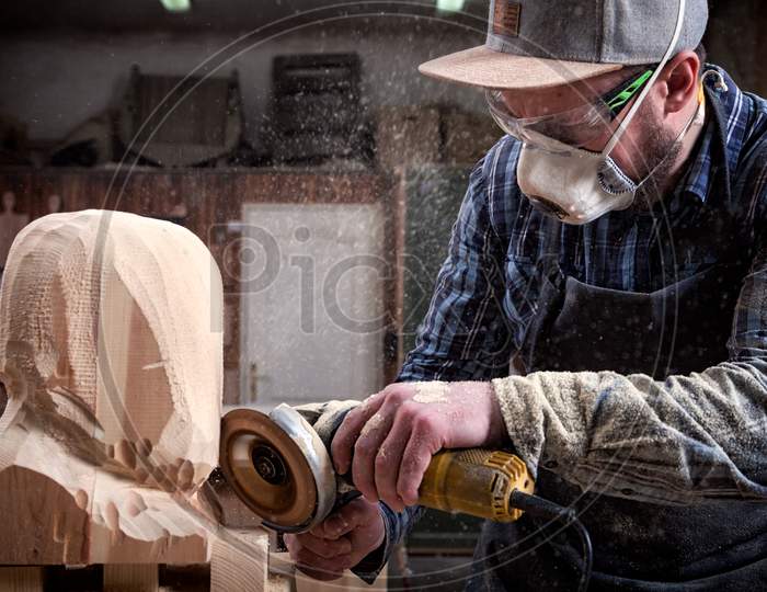 Experienced Carpenter In Work Clothes   Saws A Man'S Head With A Tree , Using An Angle Grinder  In The Workshop, Around A Lot Of Tools For Work