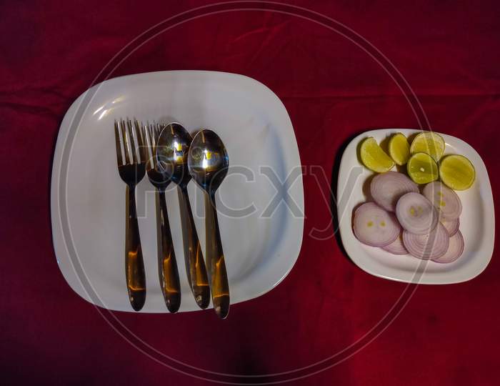 Stock Photo Of A Cut Onion Rings And Lemon With Steel Spoons And Fork Decorated On White Color Plates On Dining Table In Red Background. Captured At Night .